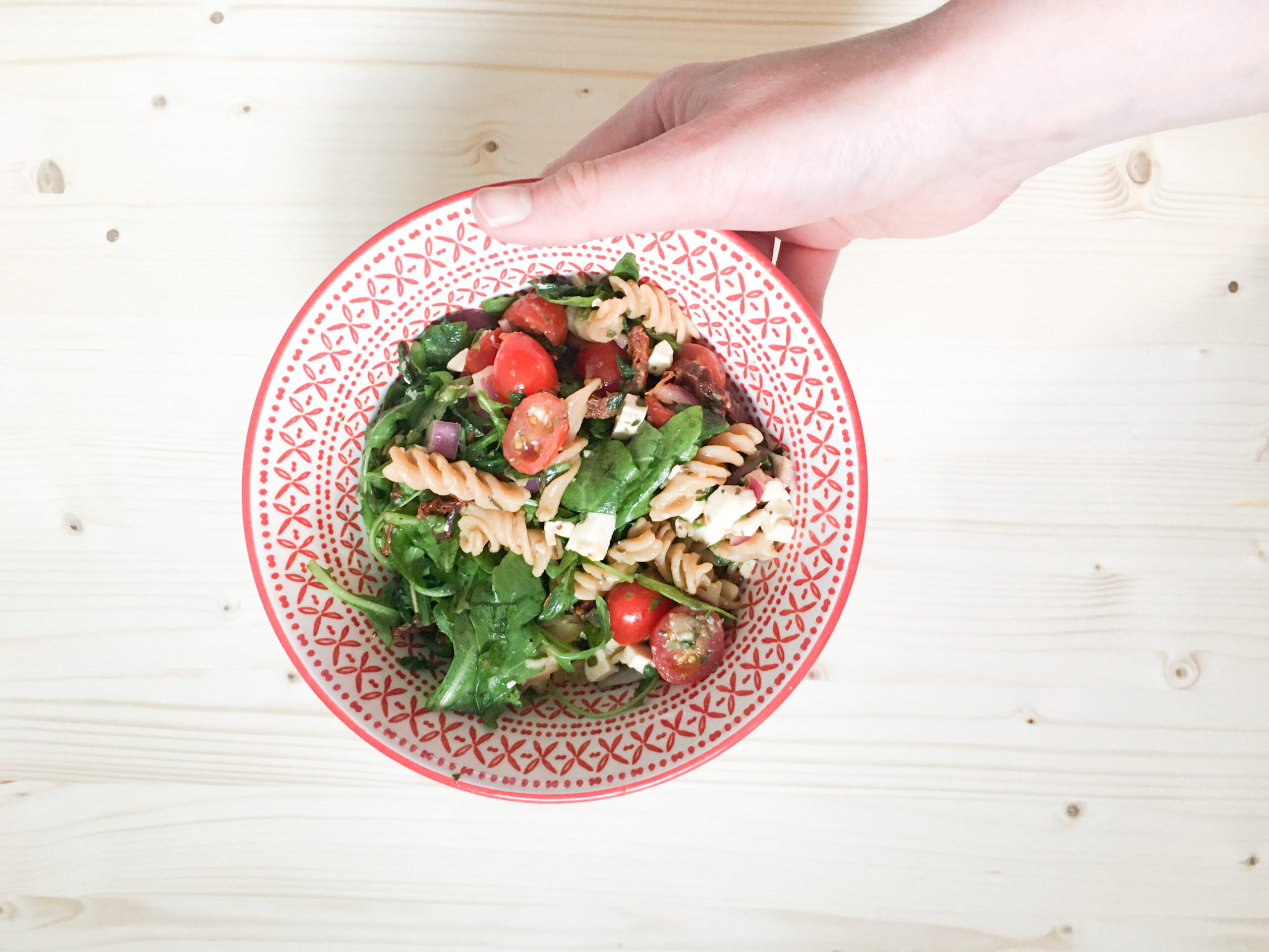 Une personne tenant un bol de salade de pâtes avec des tomates et de la roquette, sur une table en bois.