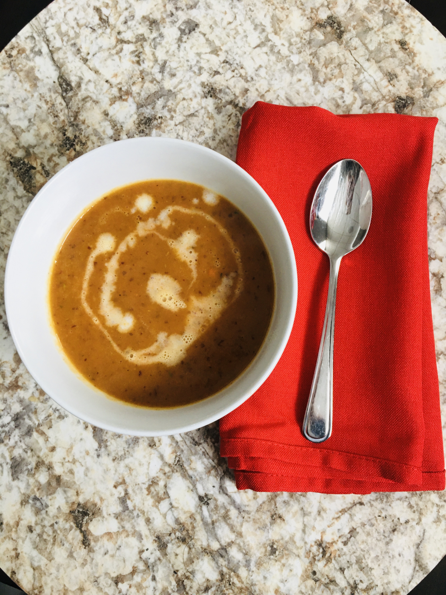 Bol de potage aux courges sur une surface en marbre, avec une serviette rouge et une cuillère en métal à côté.