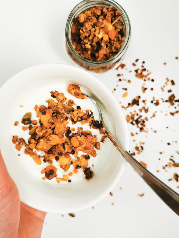 Un bol de granola maison sur du yaourt, avec une cuillère en métal, et un pot de granola en arrière-plan, sur une surface blanche.