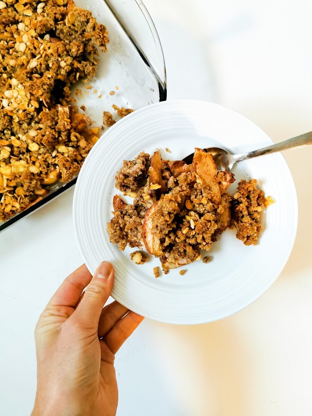Une main tient une assiette blanche avec du crumble aux pommes, près d'un plat en verre contenant la même préparation.