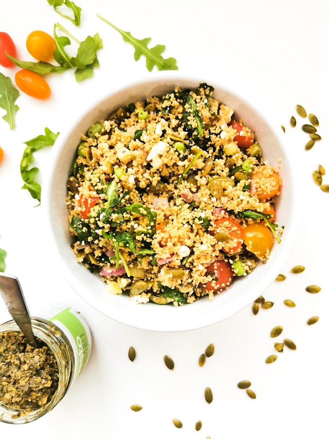Un bol de couscous coloré avec des légumes frais, des épinards, des tomates cerises et des graines, accompagné d'un pot de pesto au fond.