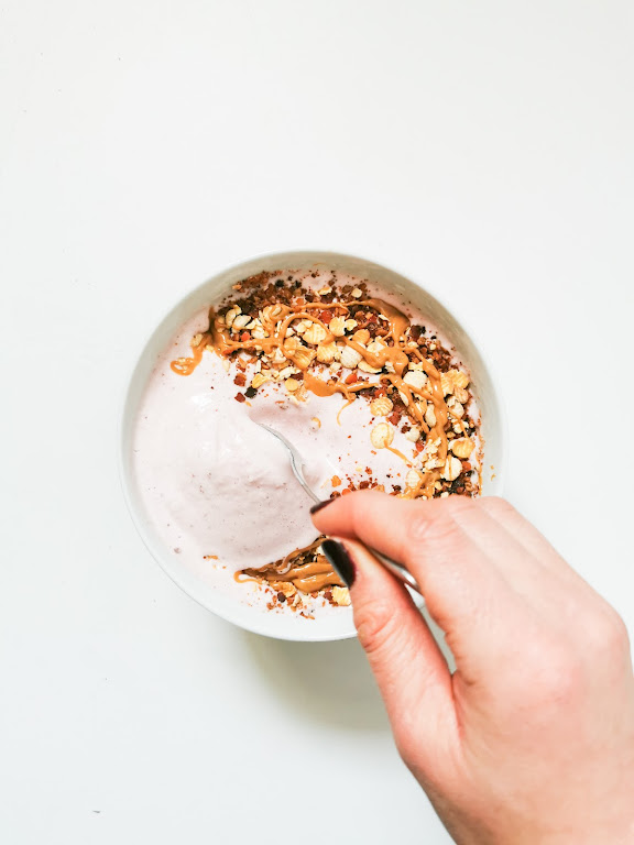 Un bol de yaourt rose avec des morceaux de noix, des éclats de céréales et une cuillère à café de beurre d'arachide, avec une main tenant la cuillère.