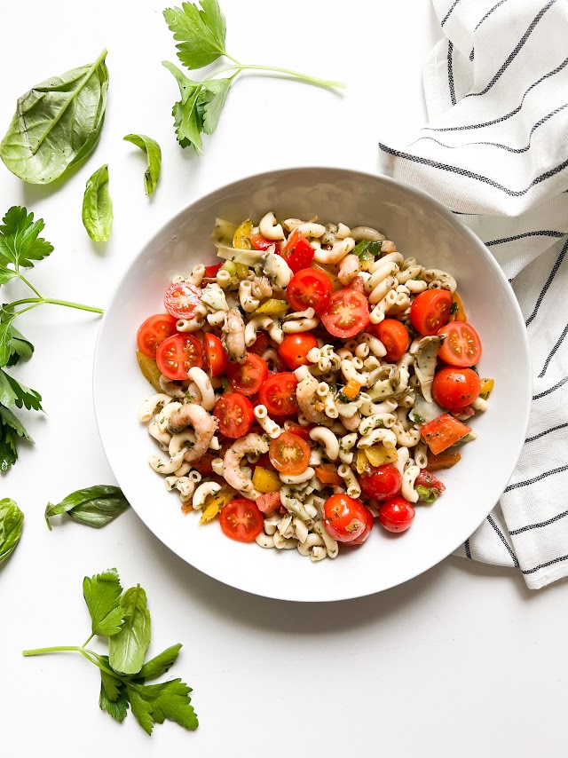 Salade de pâtes avec tomates cerises, crevettes et herbes fraîches, servie dans un bol blanc, sur une surface claire, accompagnée de feuilles de basilic et de persil.