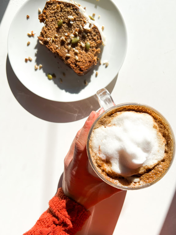 Une main tenant une tasse de café mousseux avec un morceau de pain sur une assiette à côté, sur une table lumineuse.