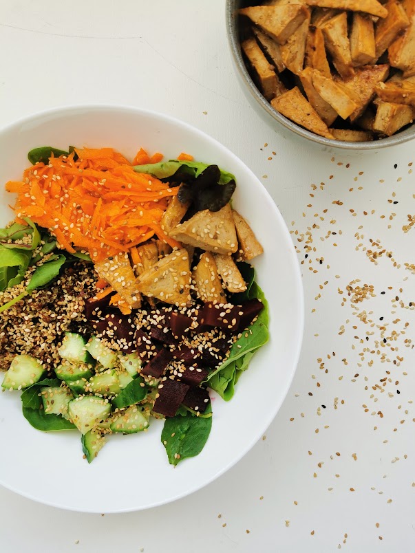 Salade colorée avec des feuilles de laitue, carottes râpées, cubes de tofu, betteraves, concombres et graines de sésame, servie dans un bol blanc.