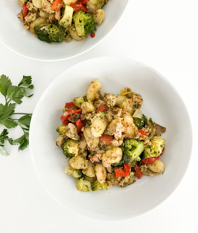 Plat de gnocchis aux légumes et poulet, garni de brocolis, poivrons rouges et aromates, servi dans un bol blanc.