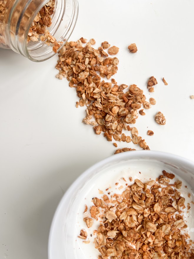 Granola croustillant versé d'un pot en verre sur un yaourt dans un bol blanc, avec des morceaux éparpillés sur la surface.