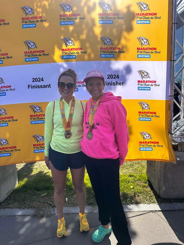 Deux coureuses célébrant leur succès à la fin d'un marathon, portant des médailles et se tenant devant un panneau de l'événement.