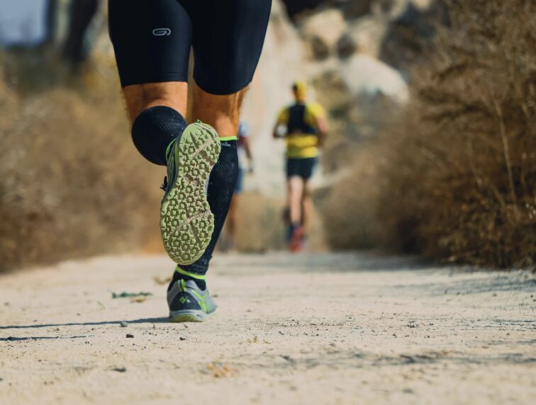 Un coureur en action sur un sentier, portant des vêtements de sport adaptés et des chaussures de course. À l'arrière-plan, deux autres coureurs sont visibles, ajoutant une dynamique sportive à la scène.