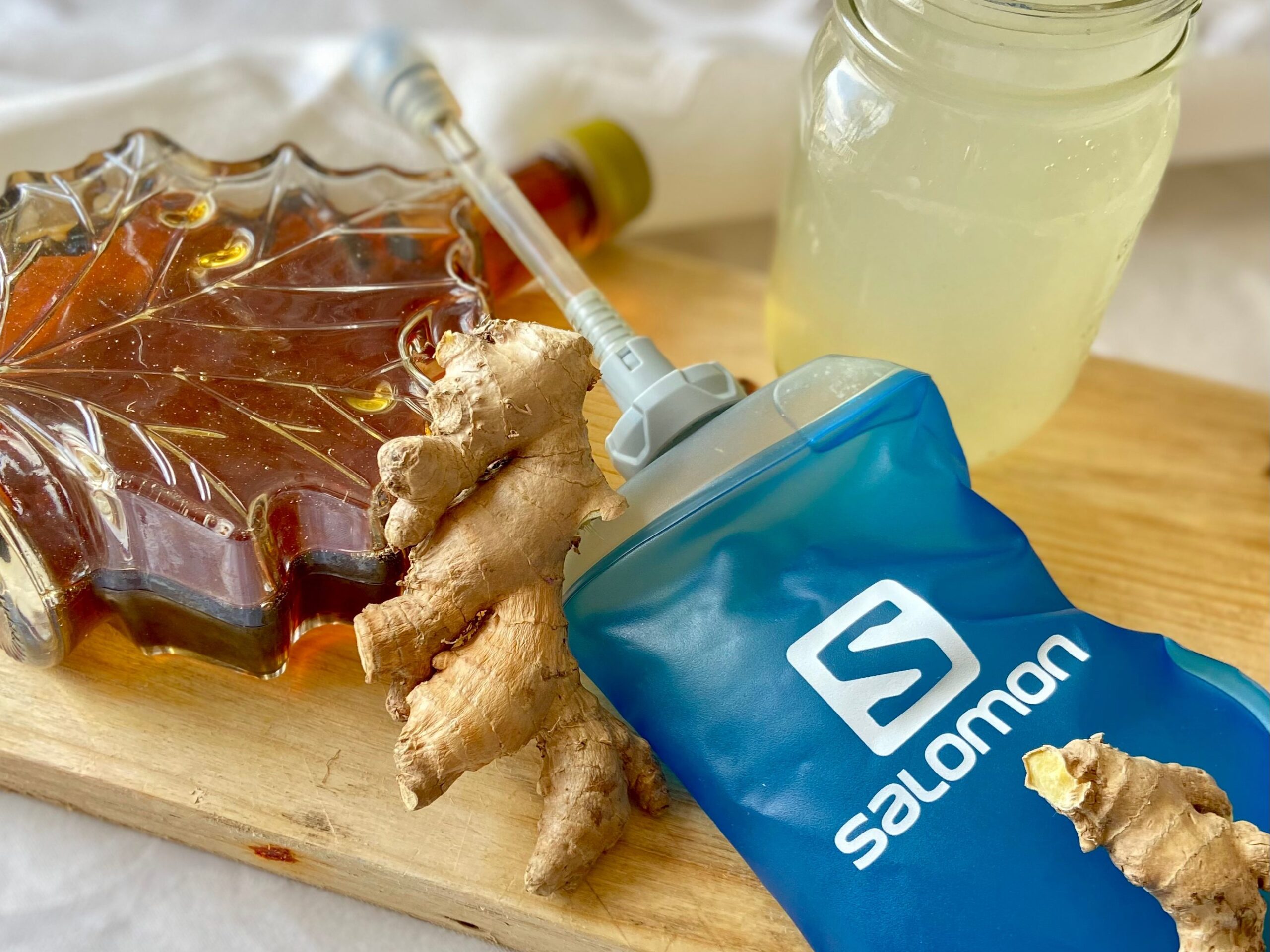 Une bouteille bleue de boisson sportive Salomon posée sur une planche en bois, à côté de racines de gingembre frais, d'un pot de sirop d'érable et d'un verre mason rempli de limonade.