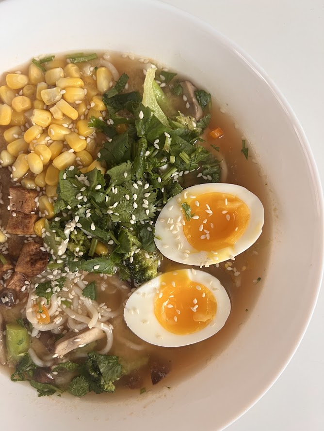 Un bol de ramen avec des nouilles, du maïs, des légumes frais, de la coriandre, des morceaux de tofu et deux œufs durs avec un jaune coulant, le tout décoré de graines de sésame.
