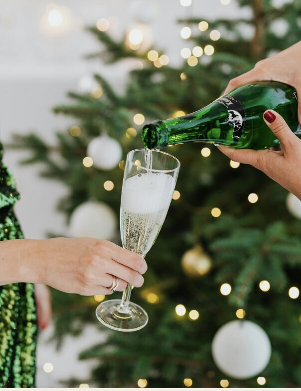 Une main verse une boisson pétillante dans une flûte, avec un sapin décoré à l'arrière-plan.