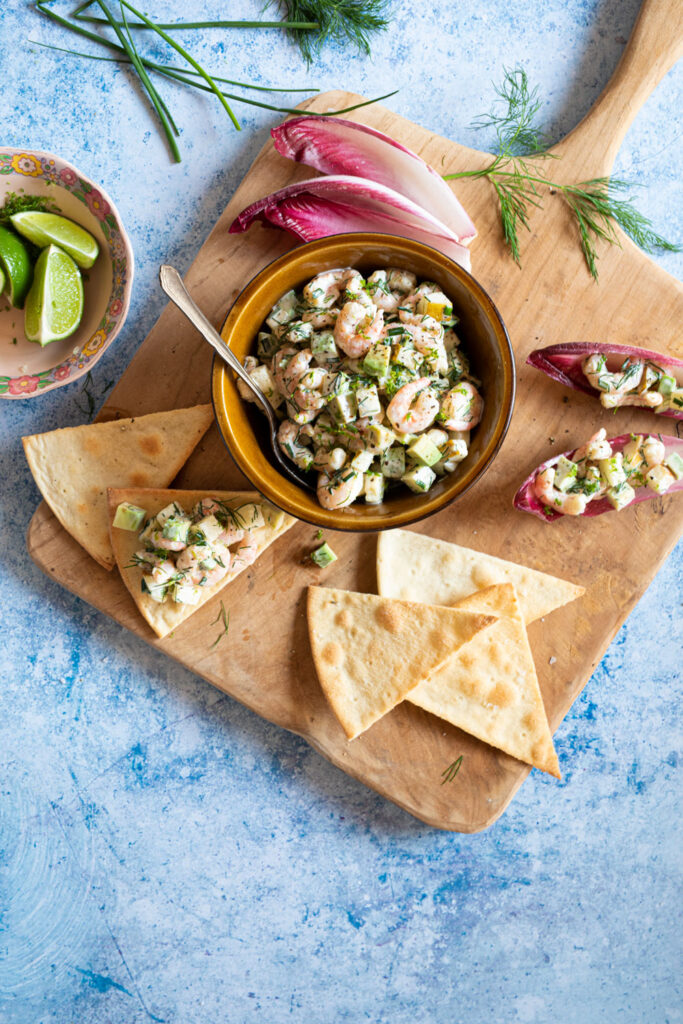 Salade de crevettes accompagnée de dés d'avocat et garnie d'herbes, servie dans un bol en terre cuite avec des chips pita et des endives. Le tout est disposé sur une planche en bois.