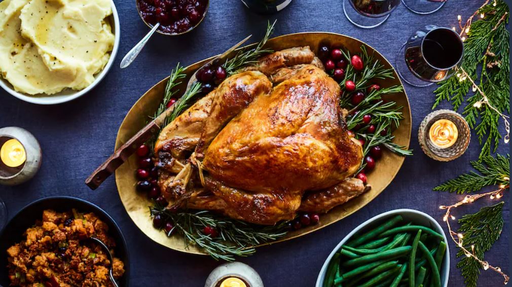 Un plat de dinde rôtie décoré avec des canneberges et des herbes, accompagné de purée de pommes de terre, de farce, de haricots verts et de sauce aux canneberges sur une table de fête.