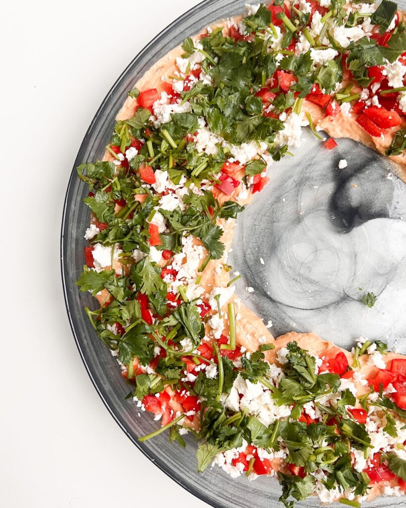 Assiette de houmous garnie de tomates coupées, fromage feta émietté et coriandre fraîche, présentée sur un plat en céramique.