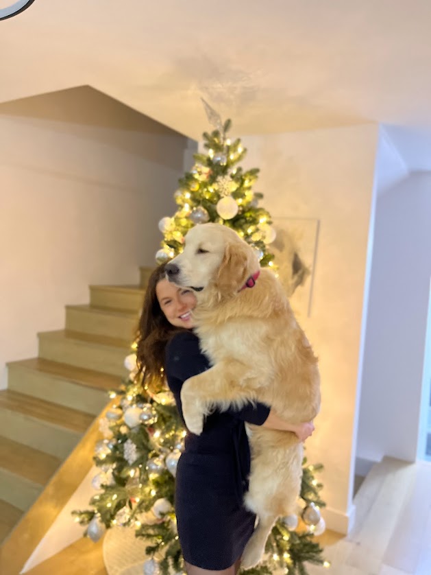 Une personne porte un chien doré dans ses bras, devant un sapin de Noël décoré.