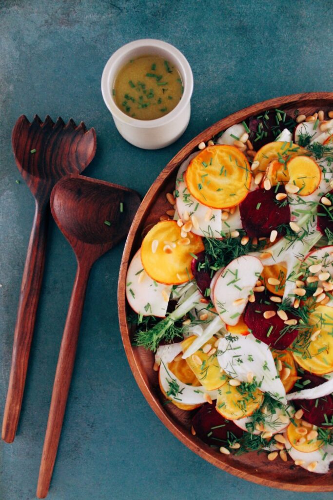 Salade colorée de betteraves, radis et légumes garnie de pignons de pin et d'aneth, accompagnée d'une vinaigrette dans un petit récipient.