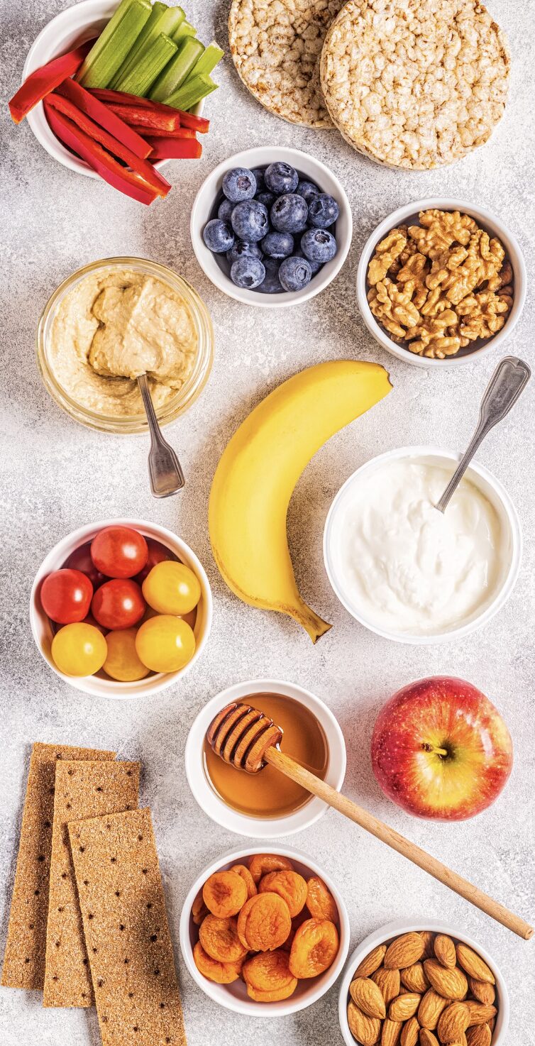 Assortiment de snacks sains comprenant des fruits frais, des noix, des légumes, du yaourt et du houmous.