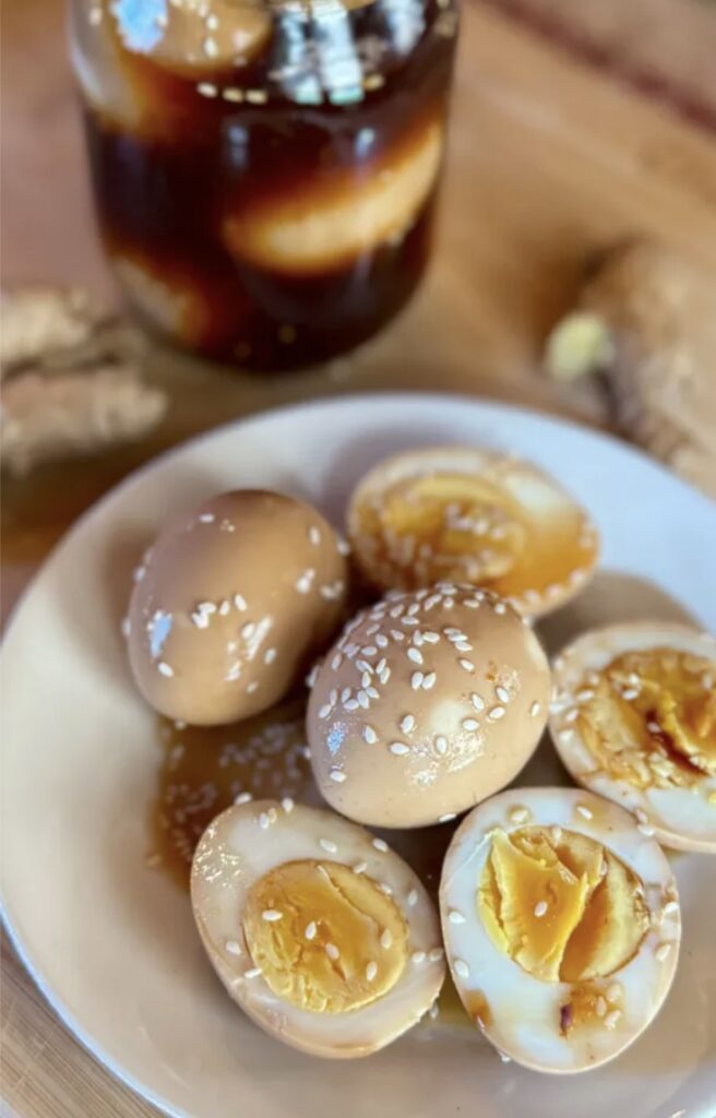 Des œufs marbrés garnis de sésame sur une assiette, avec un pot contenant des œufs dans une marinade.
