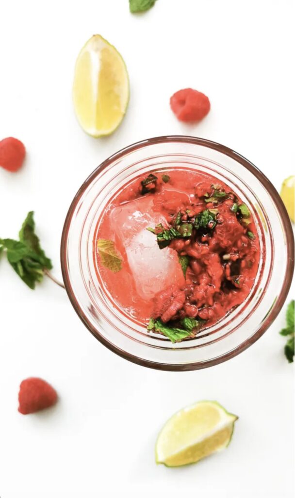 Un verre de boisson rouge avec de la menthe fraîche et des glaçons, entouré de framboises et de quartiers de citron vert.