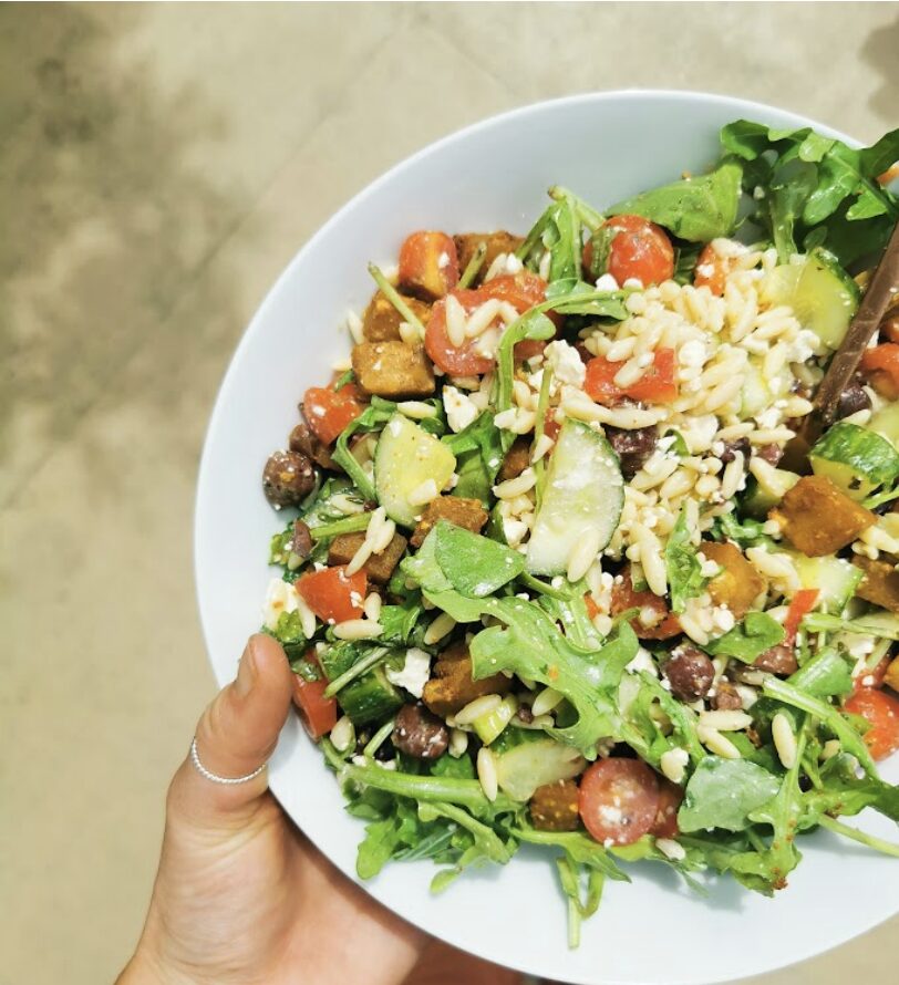 Bol de salade colorée avec roquette, tomates, concombres, grains de blé, et morceaux de légumes grillés, tenu par une main.
