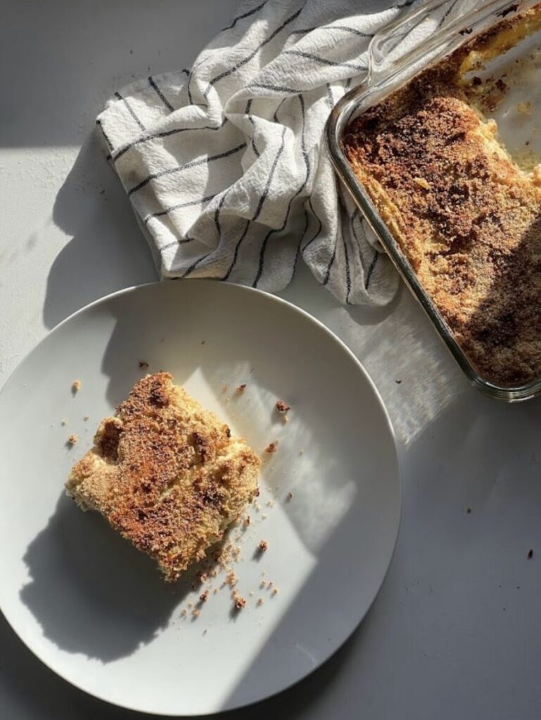 Un morceau de gâteau sur une assiette blanche, avec un plat en verre à côté. Une serviette à rayures est posée derrière, sous une lumière naturelle.