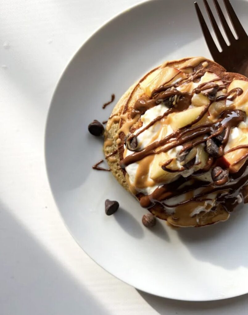 Une crêpe garnie de crème chantilly, de bananes tranchées et de sauces au chocolat et au caramel, avec des pépites de chocolat sur une assiette blanche.