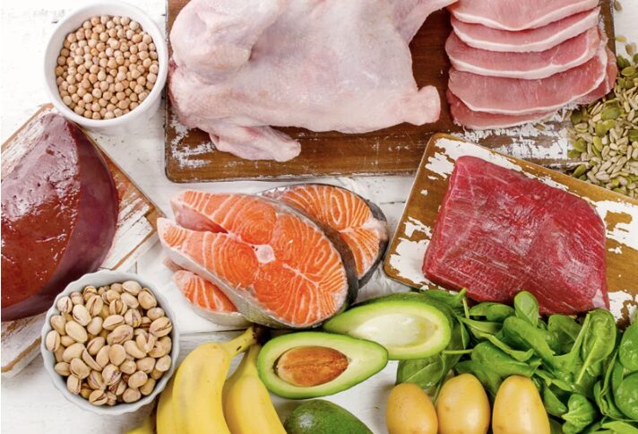 Un assortiment varié d'aliments comprenant du poulet, des tranches de viande, du saumon, des œufs, des bananes, des pistaches, des épinards et de l'avocat, disposés sur une surface en bois.