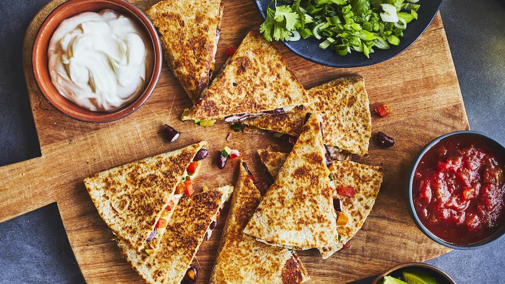 Quesadillas croustillantes garnies de légumes, servies avec de la crème, de la salsa et des herbes fraîches sur une planche en bois.