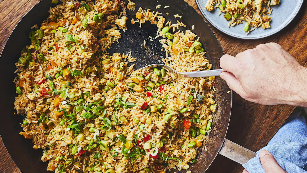 Un plat de riz sauté mélangé avec des légumes colorés, avec une main utilisant une cuillère pour servir le mélange dans une grande poêle.