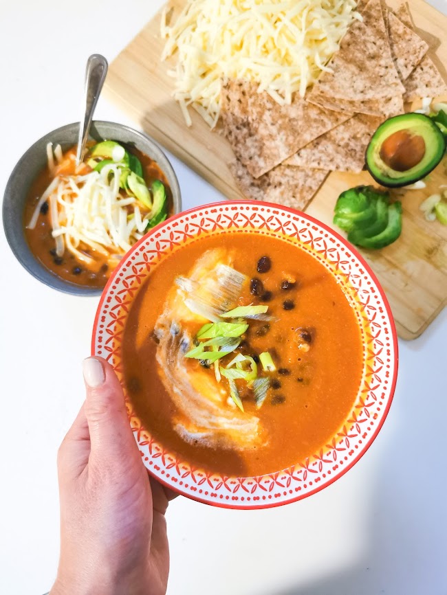 Une main tenant une assiette de soupe rouge avec des morceaux de poisson et des oignons verts, accompagnée de tortillas et d'un bol de soupe supplémentaire, avec des garnitures comme de l'avocat et des morceaux de fromage sur une planche en bois.